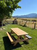 a light natural wood picnic table with attached benches sits on a green grassy lawn in a rural outdoor setting, with a fence, tree, field, horse, and mountains in the background under a clear blue sky.