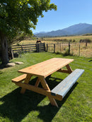 a wooden picnic table with attached benches sits on a green grassy lawn outdoors. a tree with green leaves is on the left, a wooden fence and field are in the background, and mountains are visible under a blue sky.